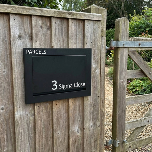 Black parcel box with 'PARCELLES' and address '3 Sigma Close' mounted on a wooden fence.  DAPRC107ANTHMATT  DAPRC107BLKMATT 